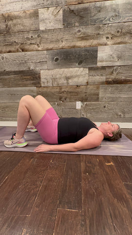 A person in pink shorts and black tank top lying on yoga mat with feet on floor and knees up