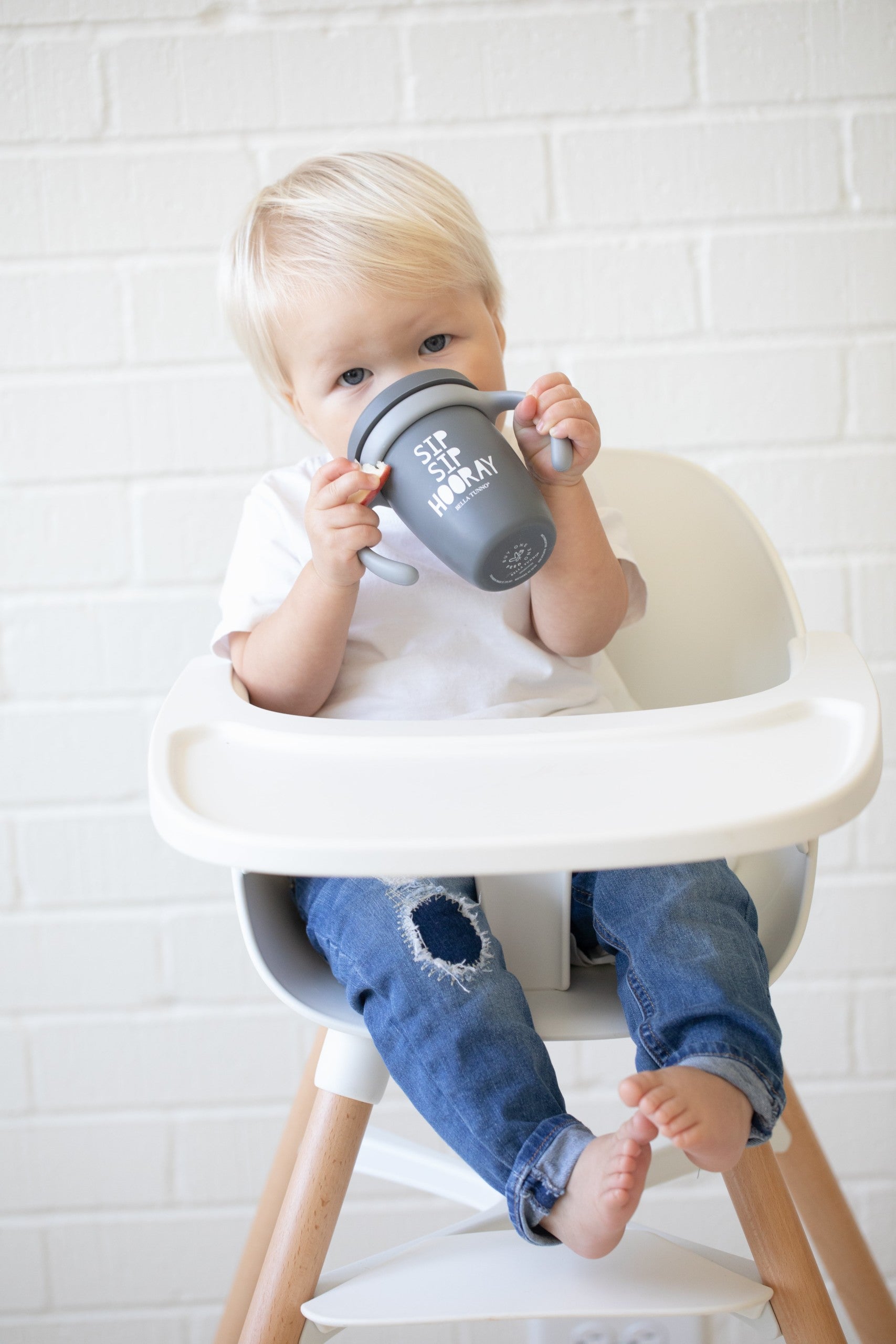 Sip Sip Hooray Happy Sippy Cup by Bella Tunno being held by a kid while in a high chair