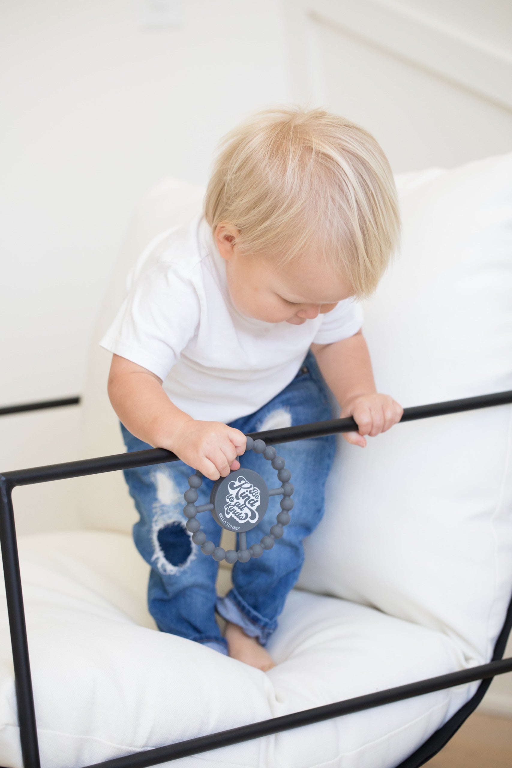 Be Kind to All Kinds Happy Teether by Bella Tunno being held by a baby boy while on the bed